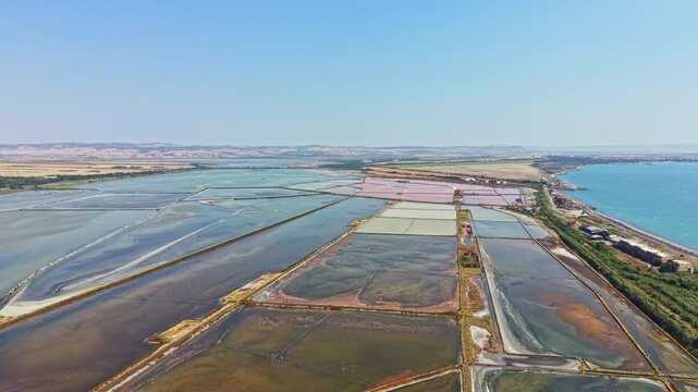 Large salt flats next to the ocean. Different sections are filled with water, creating various colors. The scene is viewed from the sky in bright sunlight.