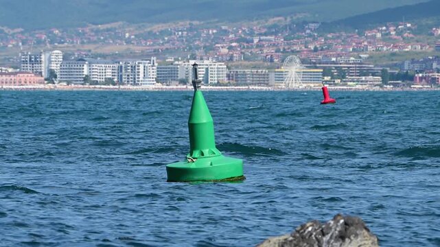A bright green navigational buoy bobs in the blue sea, with a red buoy and a coastal resort town featuring a prominent Ferris wheel in the sunny background.