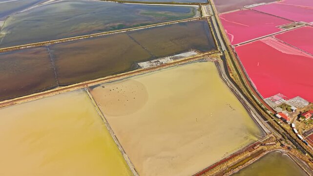 Aerial view of salt flats in a coastal region. The landscape shows patches of yellow and pink, indicating salt production. Roads and structures are present in the scene.