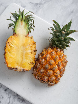 Two pineapples whole and halved on white speckled cutting board