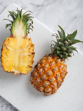 Two pineapples whole and halved on white speckled cutting board