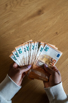 Vertical. Close-up top view of a Black woman holding and counting a large amount of fifty euro banknotes over a wooden table, representing savings, income, wealth and financial planning.