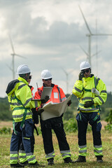 Fototapeta premium A mixed team of wind farm engineers in yellow and orange hi vis walk the meadow while consulting the site plan on a green meadow with rows of turbines behind in bright overcast daylight.