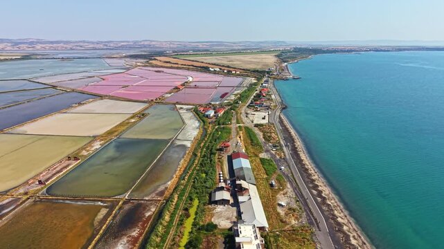 Salt farms are visible along the coast near a small town. Colorful fields are arranged in patterns beside the clear blue water. Roads and buildings are seen near the shore.