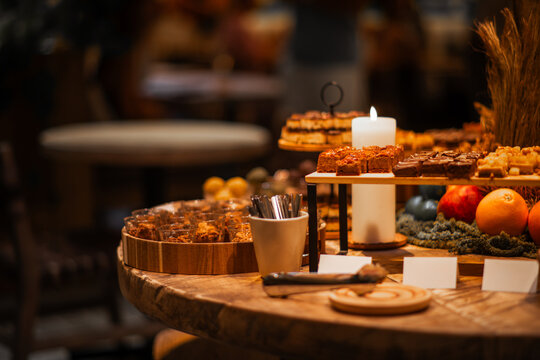 Warmly lit catering table presents handmade brownies, sliced pastries, granola cups and fresh fruit beside a glowing candle, serving utensils and blank food labels
