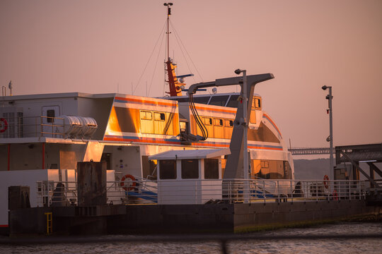 Modern passenger ferry catamaran rests at a river terminal as low evening sun casts vivid golden reflections across its windows and metal hull, with a distant bridge beyond