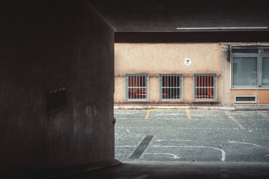 Shadowy covered entrance frames a vacant paved inner yard with marked parking bays, barred windows and a weathered industrial facade glowing beyond the tunnel