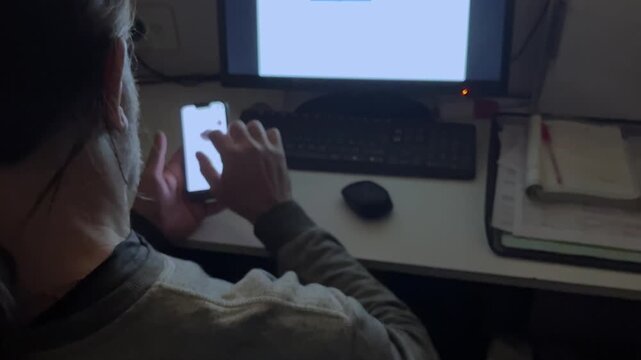 Concentrated young adult sitting at desk in dark room scrolling and tapping on illuminated smartphone screen with computer monitor and keyboard in the background