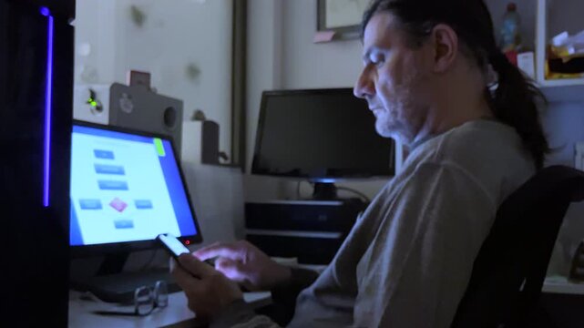 A man sits intently at a desk in a dimly lit room, focused on his computer and looking at information on his smartphone.