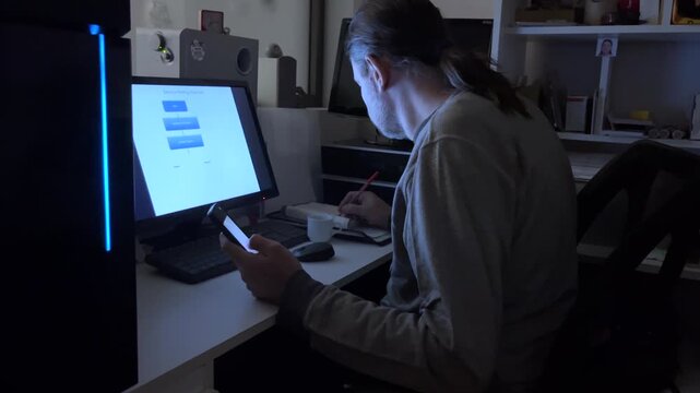 Mature adult man using his smartphone and computer in a dark room illuminated by the screens while the computer case shows a variable spectrum led light display