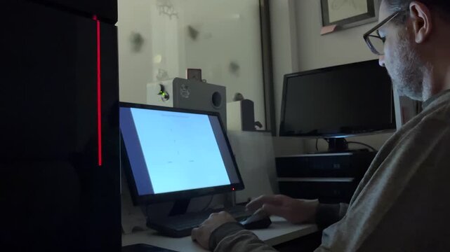 Mature man working on a computer creating a flowchart in a dark room, his face illuminated by the screen with a gaming pc with colorful changing led lights