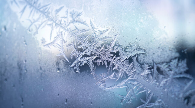 Close-up of intricate ice crystals and frost patterns spreading across a cold glass window pane with soft blue light and frozen condensation droplets in winter.