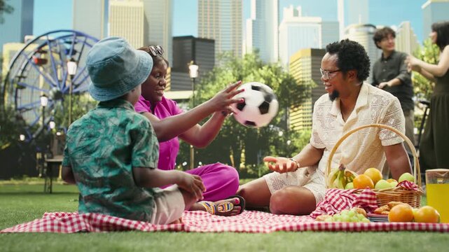 Parents laugh and play ball game with jolly boy on picnic blanket, sharing active summer afternoon in green urban park with picnic arrangement and food.
