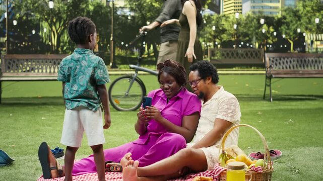 Mother photographs her smiling boy with a smartphone in portrait orientation while capturing adorable moments during a picnic in a city park.