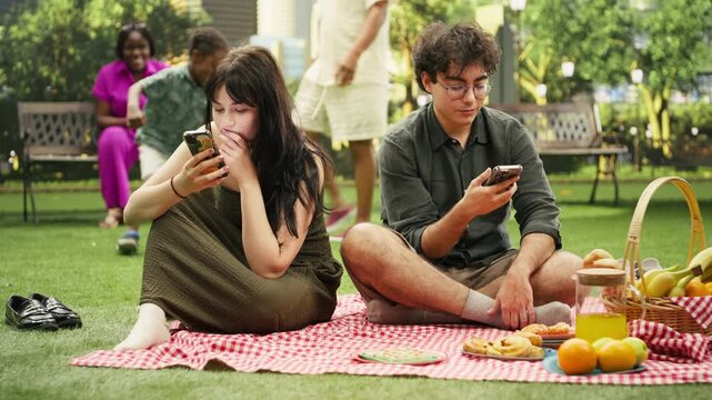 Disconnected couple scroll through social media during a picnic date, showing digital distraction, silence and disengagement between them.
