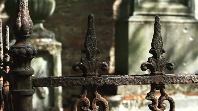 Long pan across an old, rusted wrought iron cemetery gate with ornate details and pointed spikes. Neutral tones and natural light highlight the intricate, weathered texture and underlying tombs.
