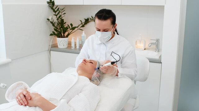 Masked beautician uses microcurrent electrodes on Caucasian woman face during anti aging cosmetic treatment in clinic. Client lies relaxed on bed receiving electrical impulse facial therapy session.
