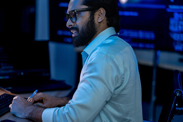 Fototapeta premium South Asian male tech lead in a suit types at a keyboard while multiple screens display network security dashboards in a dark office.