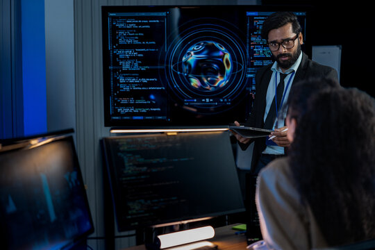 South Asian male engineer instructs a Black female analyst on security protocols in a dark tech office with glowing monitors.