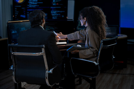 South Asian male and Black female software developers pair-program at dual monitors displaying network security algorithms at night.