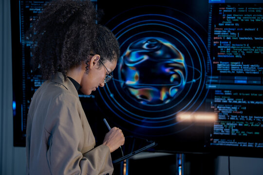 Black female data analyst cross-references code on a monitor with notes on a tablet in a blue-lit nighttime operations center.