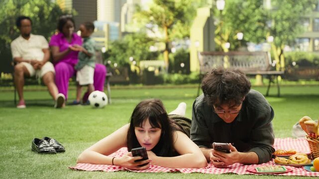 Disconnected couple scroll through social media while lying on a picnic blanket, showing digital distraction, phone dependency and disengagement between them.