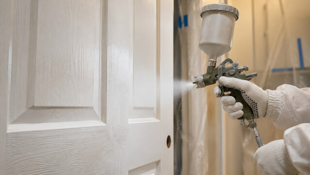 A person wearing gloves and protective clothing spray paints a white wooden door using a paint spray gun in a controlled indoor environment.