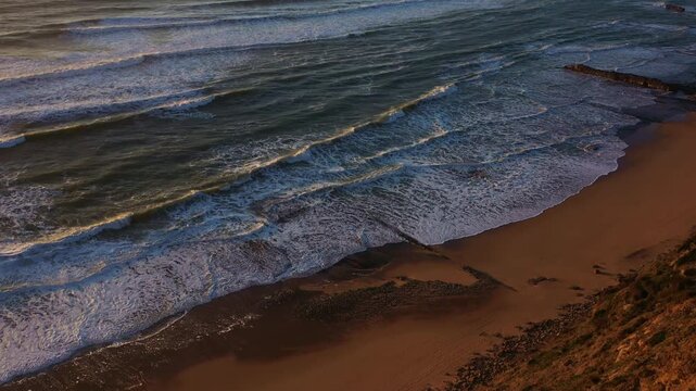 Waves roll on the sandy beach as the sun sets in a coastal area aerial view. The sky shows different shades as the light changes over the water and sand.