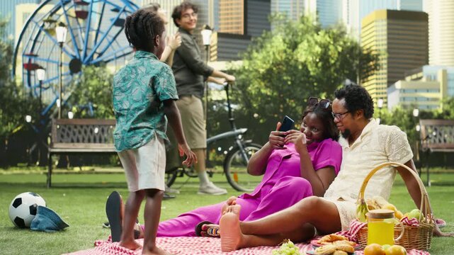 Proud mother takes pictures and records the boy using a mobile phone during a picnic in a city park, capturing an adorable family moment and photographing her young son during family time.
