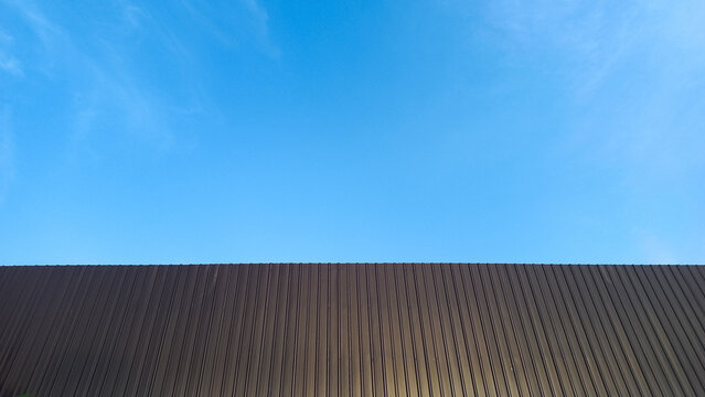Minimalist view of a dark brown corrugated metal roof against a clear bright blue sky. Clean horizontal line dividing the roof and sky. Industrial building exterior with simple geometric composition, 