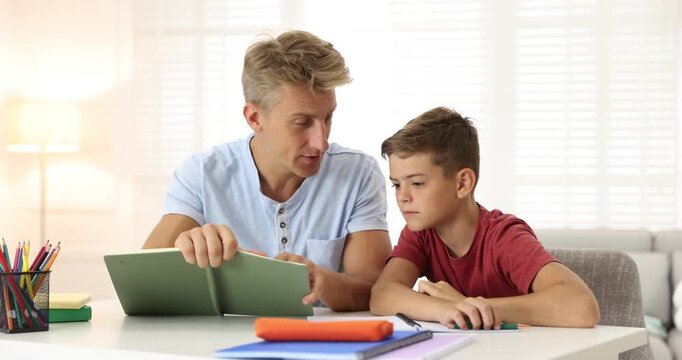 Father with book helping his son with homework at white table indoors