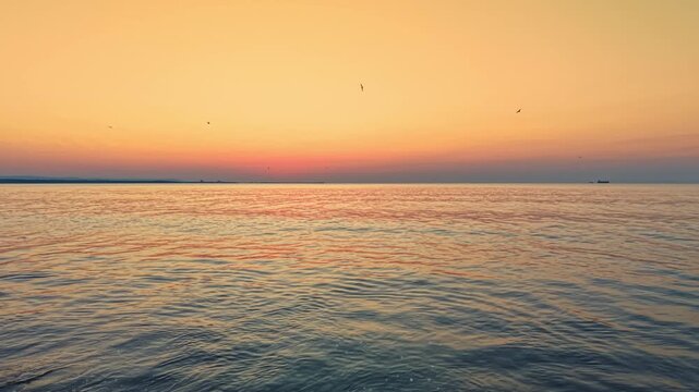 The ocean reflects the colors of the sunset as waves gently move towards the shore. Boats can be seen on the horizon during this evening scene.