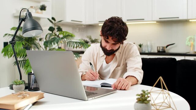 Indian man at home listens to online lecture on laptop writes notes in notebook feels focused. Arabian guy sitting at table looks at screen turns pages marks points in copybook feels study motivation.