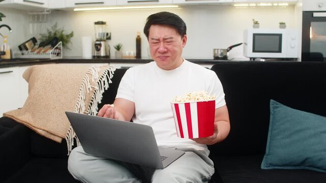 Asian man at home watches comedy on laptop eats popcorn and laughs out loud enjoys jokes and fun. Chinese guy on sofa smiles wide repeats funny line takes popcorn and relaxes while movie keeps going