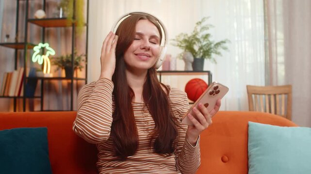 Young woman at home uses wireless headphones starts playlist nods to beat smiles and types reply on smartphone. Caucasian girl on sofa listens to music and chats by message feels happy, chill, relax