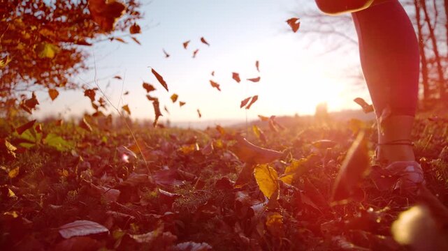 LENS FLARE, LOW ANGLE, CLOSE UP, DOF, SLOW MOTION: Woman running through forest at sunset, kicking up dry autumn leaves into the air. Energetic outdoor activity in the swirl of flying fall foliage.