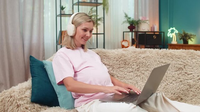 Pregnant middle-aged woman at home wears headphones editing documents on laptop working freelance feeling calm. Caucasian girl on sofa enjoys music finishing tasks joyful calm soft evening mood.