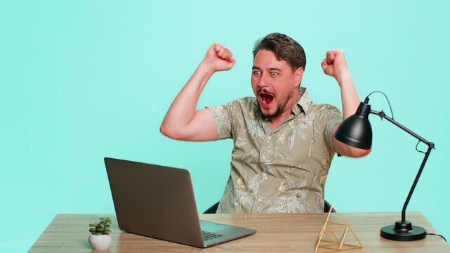 Middle-aged man sits at table, joyfully raising both arms in celebration after successfully completing an online course or exam on laptop. Young adult guy on blue background expressing achievement.