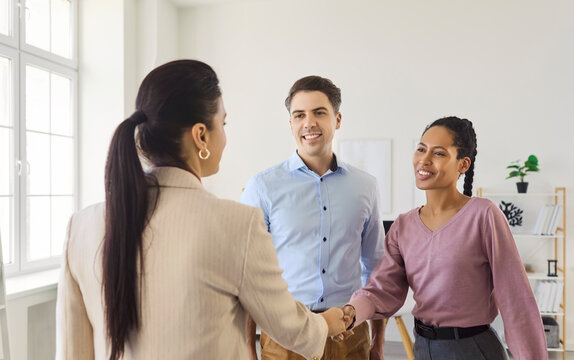 Agent and couple handshake at property meeting. Friendly realtor welcomes clients in a flat during a consultation, discussing rental agreement and contract details. Trust and deal theme shown.