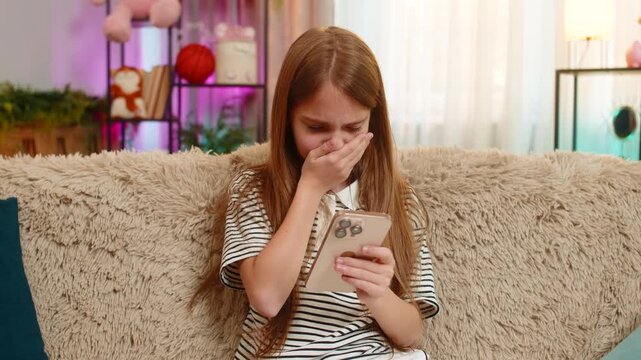 Young child girl sits on sofa and looks sadly at smartphone after losing mobile game or reading bad school test result. School kid shows frustration disappointment, emotional reaction to negative news