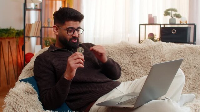 Indian man at home reacts to Bitcoin BTC drop, closing laptop and holding coin with unhappy look. Arabian guy on sofa expresses disappointment and stress, emotionally affected by falling crypto value