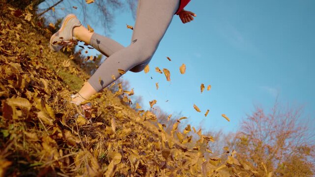 CLOSE UP, LOW ANGLE, SLOW MOTION: Woman jogging along a slope as autumn wind swirls fallen dry leaves. Motion, sport and the playful energy of an outdoor workout in a sunny countryside setting in fall