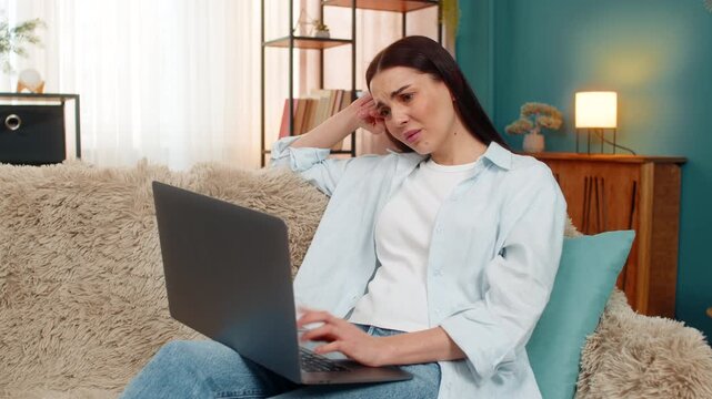 Young woman at home dissatisfied looking at laptop screen leaning head on hand tiredly with upset expression. Caucasian girl on sofa feeling fatigue, frustrated from workload and negative emotions