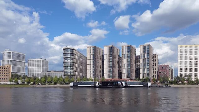 Timelapse of a modern residential quarter on the Moskva River embankment in Moscow. Electric river buses call at the floating pier. Cumulus clouds drift over the new apartment blocks all day.