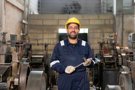 Confident technician wearing hardhat smiling while holding wrench surrounded by machinery in factory.