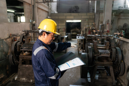 Technician wearing hardhat reading and inspecting report folder surrounded by machinery in factory.