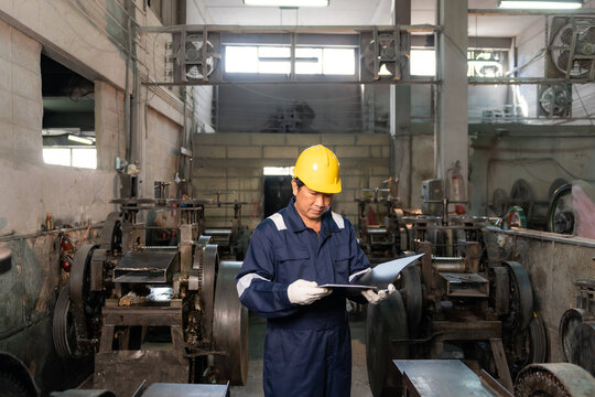 Technician wearing hardhat reading and inspecting report folder surrounded by machinery in factory.