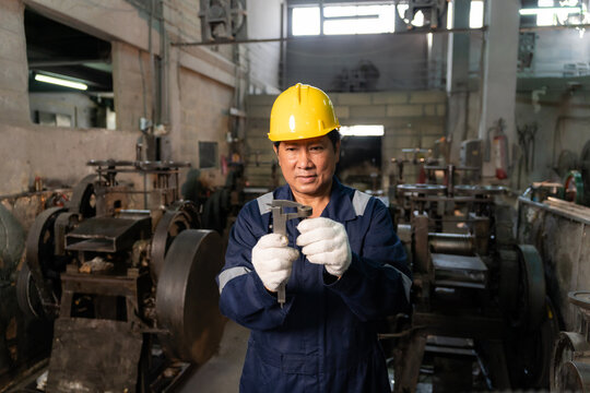 Confident technician wearing hardhat using vernier caliper to measure and inspect metal component in factory.