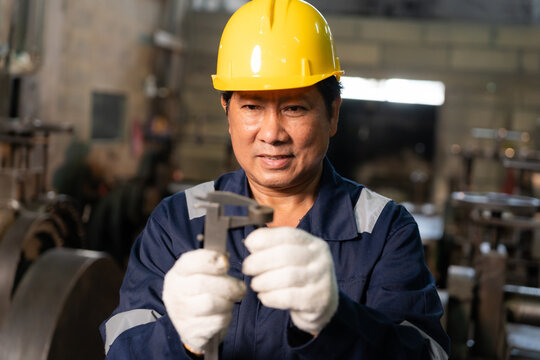Confident technician wearing hardhat using vernier caliper to measure and inspect metal component in factory.