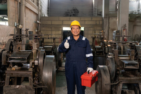 Confident technician wearing hardhat showing thumbs up and holding toolbox in industrial factory.